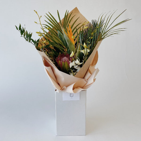 A tropical flower bouquet with palm leaves, orchids, protea and birds of paradise flower displayed in an upright delivery box and wrapped in kraft paper