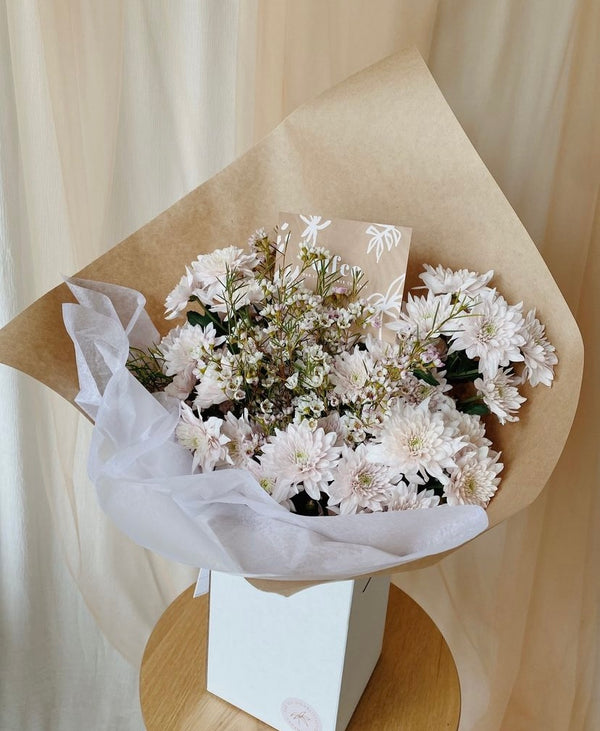 Light pink spray mums with wax flower filler displayed in an upright delivery box and wrapped in kraft paper